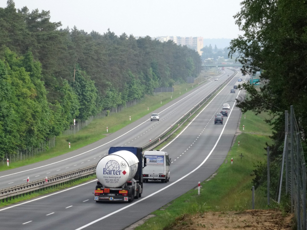 Ein großer Lkw fährt auf einer von beiden Seiten von Bäumen gesäumten Autobahn, mit einem Zaun und Gras auf der rechten Seite und Gebäuden im Hintergrund unter einem klaren blauen Himmel.