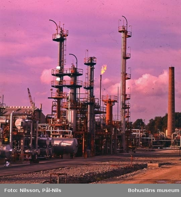 Eine Ölfabrik mit zahlreichen Rohren, Ventilen, Metallkonstruktionen, Containern, einem Kran, einem Gebäude und einer Gruppe von Bäumen unter einem bewölkten Himmel, mit Text unten.