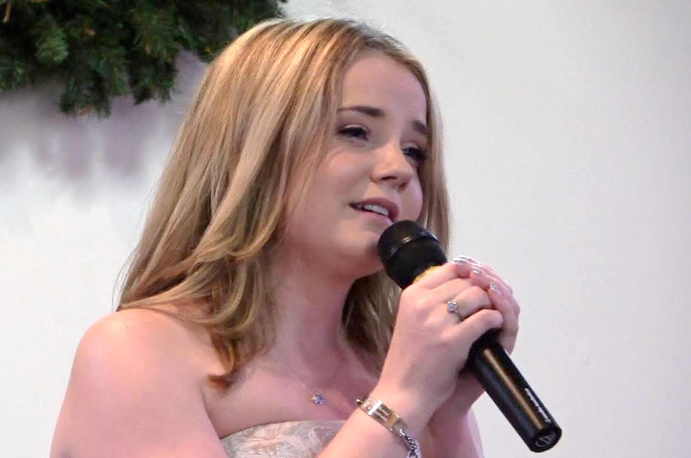Eine Frau in einem weißen Kleid singt in ein Mikrofon mit einer Wand und verlässt die Bühne im Hintergrund.