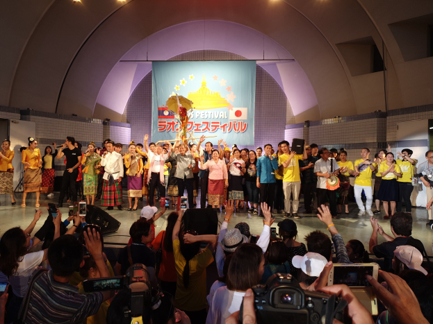 Eine Gruppe von Menschen auf einer Bühne mit Kameras, Handys und Musikinstrumenten während einer scheinbar festivalartigen Eröffnungszeremonie, mit Bannern, Wänden und Deckenlampen im Hintergrund.