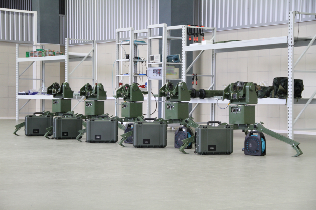 Eine Gruppe von Militärrobotern ist auf einem Boden positioniert und von Koffern und anderen Gegenständen auf Gestellen umgeben, mit einer Wand und einem Geländer im Hintergrund.