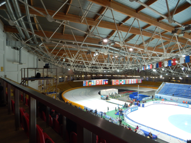 Große Indoor-Eislaufhalle mit zahlreichen Menschen, die Schlittschuh laufen, umgeben von Sitzplätzen, Geländern, Brettern, Leuchten und strukturellen Elementen, mit einer Wand im Hintergrund.