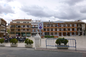 Ein belebter Stadtplatz mit Menschen auf Stühlen und Stehenden, umgeben von Topfpflanzen, Metallabsperrungen, Straßenlaternen mit Fahnen, einem Schild, Gebäuden mit Fenstern und einem bewölkten Himmel.