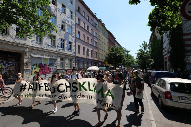 Eine Gruppe von Menschen marschiert auf einer Straße mit einem Banner, mit parkenden Autos auf der rechten Seite, einem Fahrradfahrer auf der linken Seite, Gebäuden und Bäumen im Hintergrund und einer Berliner Demonstration.