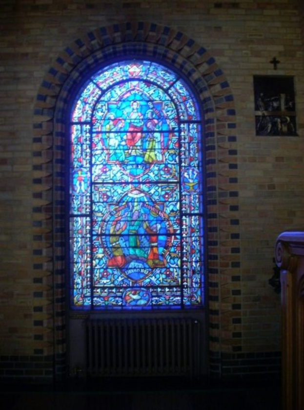 Ein Innenblick in eine Kirche mit einem heiligen Kreuzsymbol an der Wand, Buntglasfenstern, einem dunklen unteren Bereich und einem hölzernen Objekt auf der rechten Seite.